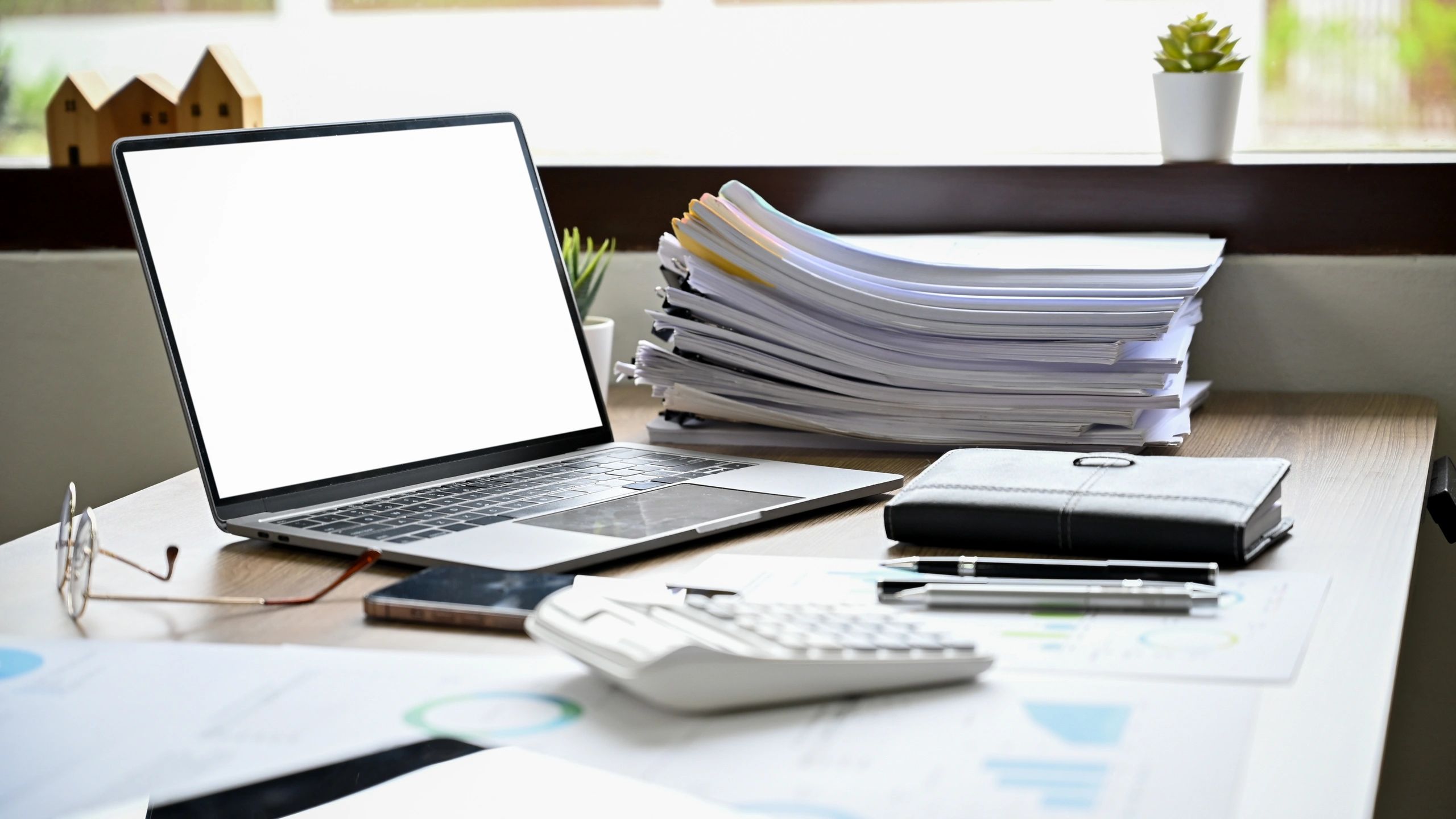A cluttered office desk with a laptop, papers, calculator, and plants.