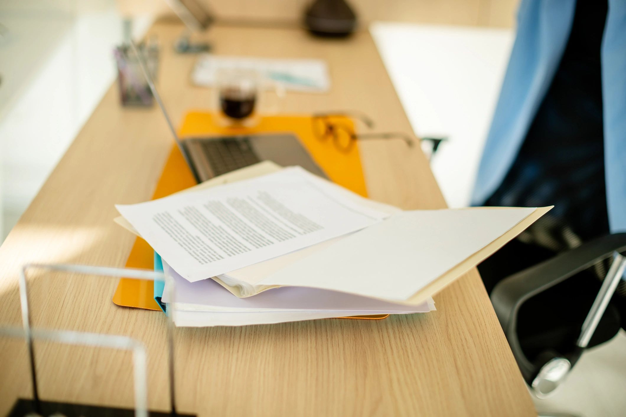 Documents and laptop on a wooden desk with a glass of coffee and glasses.